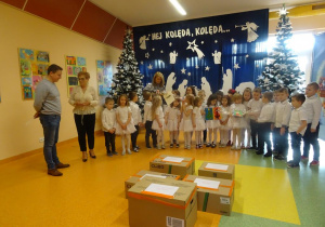 Pani dyrektor Maria Królikowska przekazuje panu Zielińskiemu życzenia świąteczne dla mieszańców hospicjum, Dzieci trzymają kartki świąteczne, na środku paczki z darami.